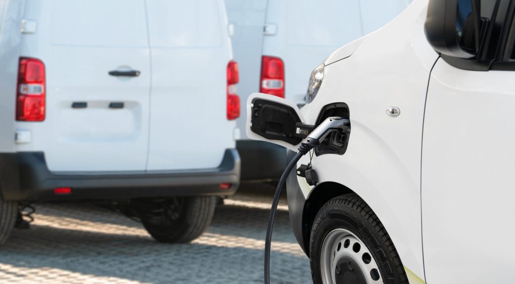Flotte von Lieferwagen mit E‑Van an Ladestation im Außenbereich