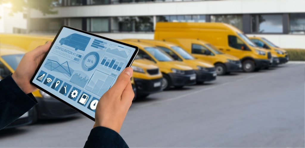Person hält ein Tablet mit Telematik-Dashboard zur Fahrzeugüberwachung, im Hintergrund steht eine Reihe gelber Lieferfahrzeuge auf einem Firmenparkplatz.