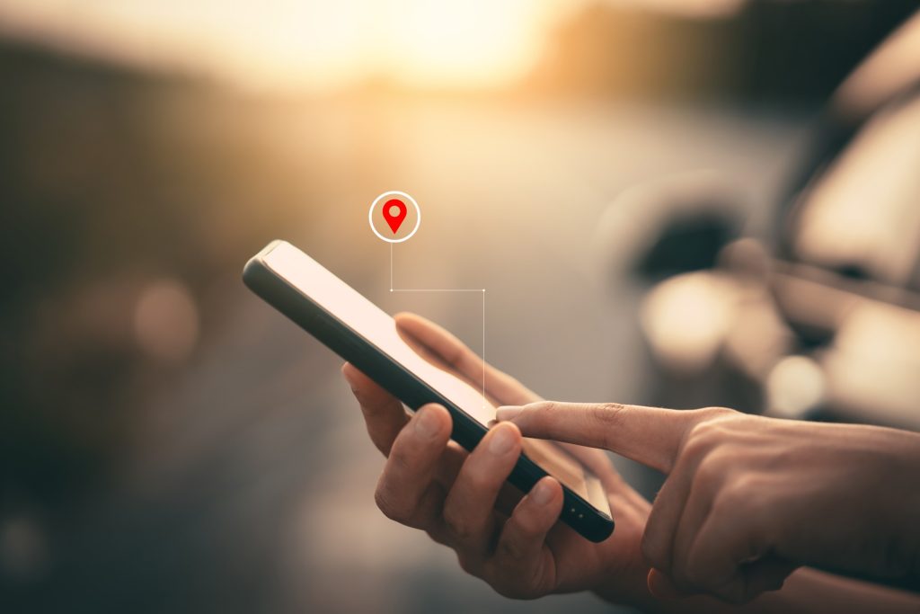 Hand hält ein Smartphone mit Navigationsanzeige; rote Standortmarkierungen und eine gepunktete Route symbolisieren GPS-Navigation und Routenplanung.