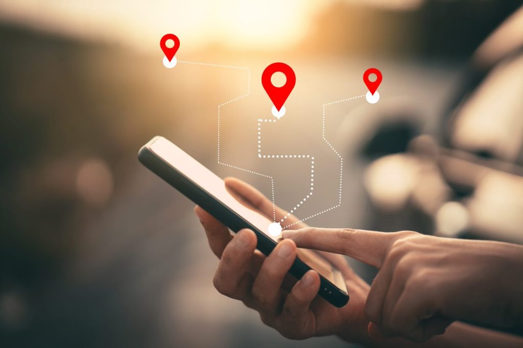 Hand hält ein Smartphone mit Navigationsanzeige; rote Standortmarkierungen und eine gepunktete Route symbolisieren GPS-Navigation und Routenplanung.
