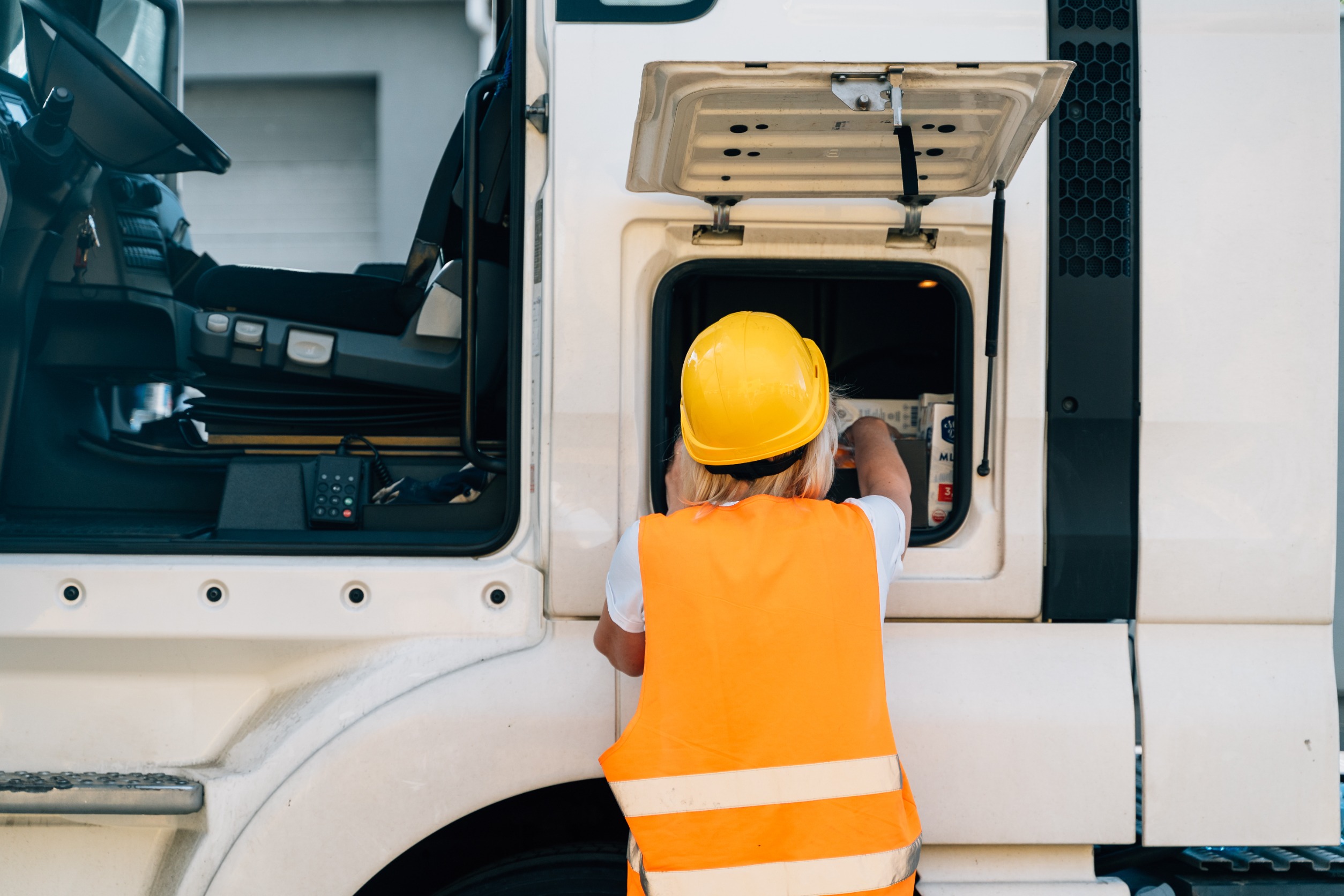 Person mit gelbem Schutzhelm und orangefarbener Warnweste überprüft ein offenes Wartungsfach an der Seite eines LKWs.